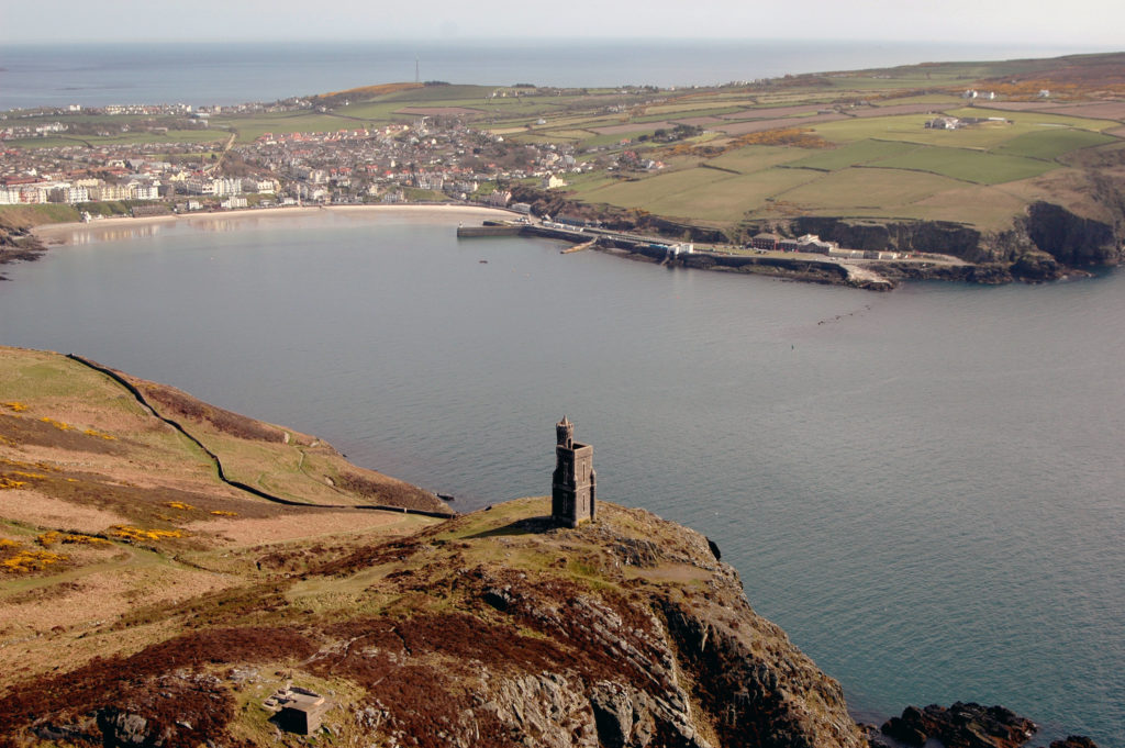 An aerial view of Port Erin Bay taken from a plane, looking towards an open clear sunny bay, with Port Erin's landmark "Milner's Tower" almost below the plane, in the distance is the nearby town of Port St Mary, and beyond that the horizon on the farther side of the island.